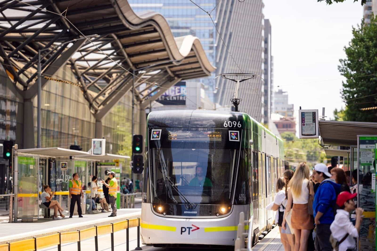 ヤラ・トラムの緑と白の E-Class Bombardier 6096「PT」が Casino / Melbourne Convention and Exhibition Centre の停留所に入線。運転台上部に白枠の NaviLens コードが 2 つ。右側では、デジタル発車案内「12a · 96 · St Kilda Beach」の前で乗客が待っており、背後にうねるような展示会場の屋根
