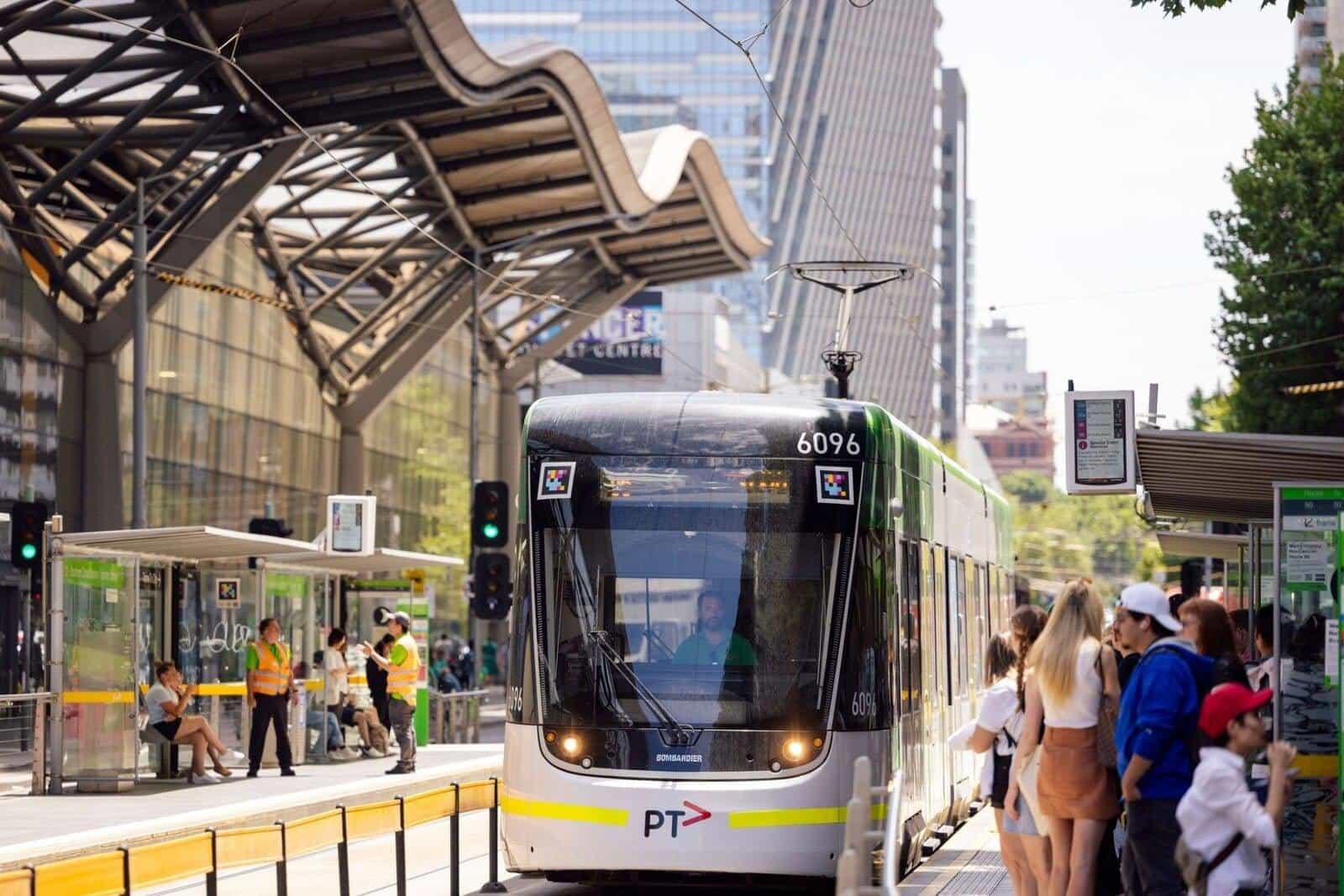 Yarra Trams green-and-white E-Class Bombardier tram 6096 “PT” pulling into the Casino / Melbourne Convention and Exhibition Center stop, with two white-framed NaviLens codes above the cab and, on the right, passengers waiting next to the digital next-departure board “12a · 96 · St Kilda Beach”; the wavy roof of the exhibition centre behind