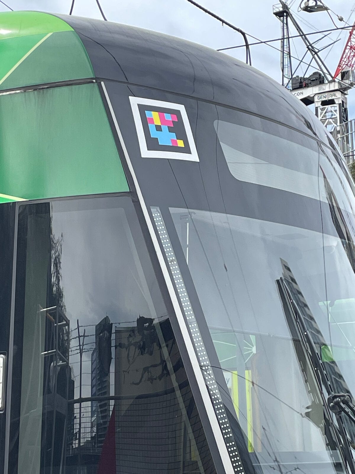 Close-up of the nose of a green-and-grey Yarra Trams E-Class tram: on the front, over the dark side window, a white-framed NaviLens code next to a decorative perforated band; the cranes and high-rises of central Melbourne reflected in the background