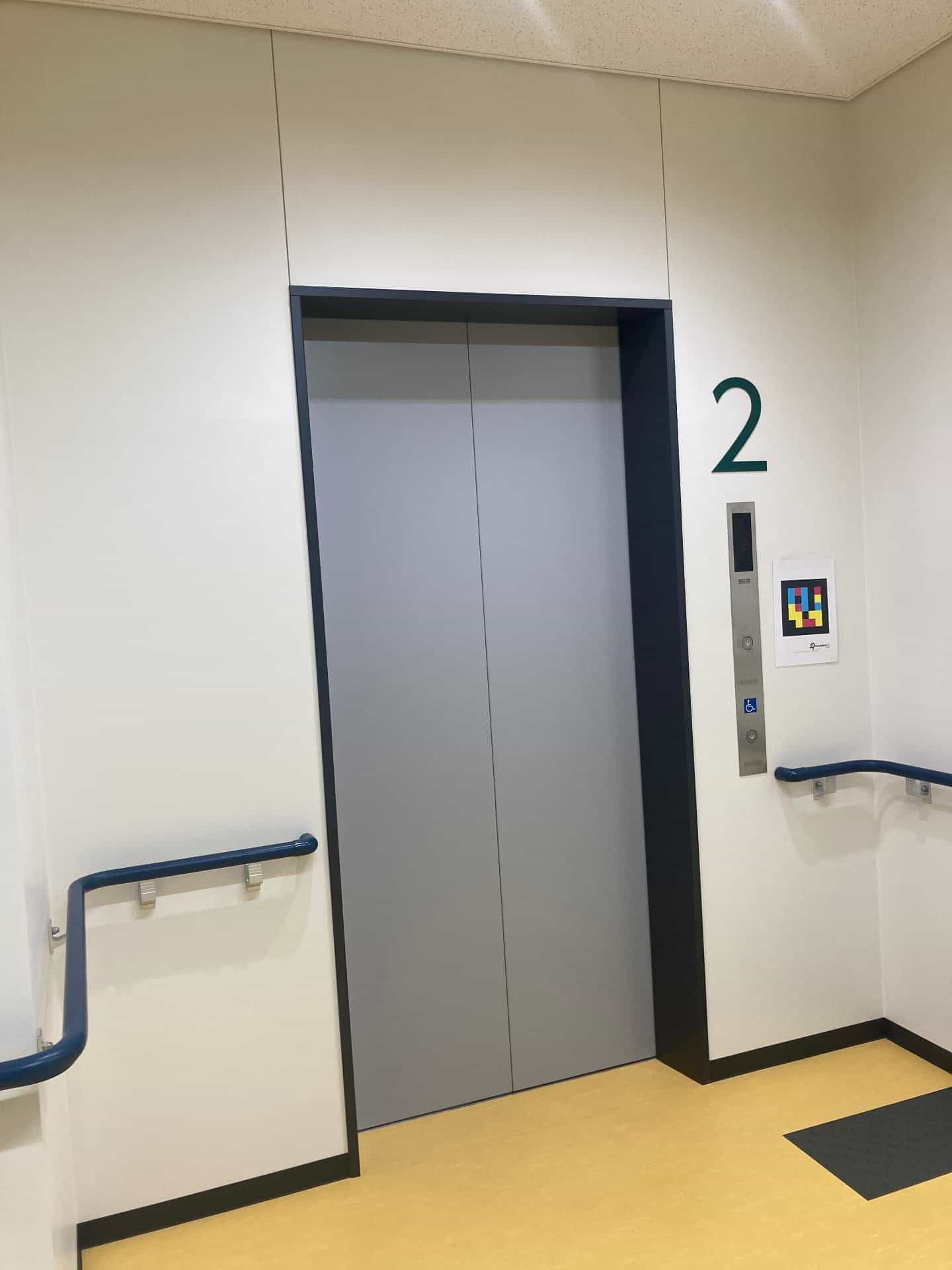 Atrio del 2º piano del Miyagi Information Center for the Visually Impaired a Sendai: ascensore con porte grigie incassate in una parete bianca e un grande «2» verde dipinto a destra; sopra la pulsantiera in acciaio inox e accanto al pulsante con l’icona della carrozzina, un cartello A4 con il codice NaviLens GO in ciano, magenta e giallo su fondo nero; corrimano tubolari blu su entrambi i lati, pavimento in vinile giallo e tappetino nero antiscivolo davanti all’ascensore