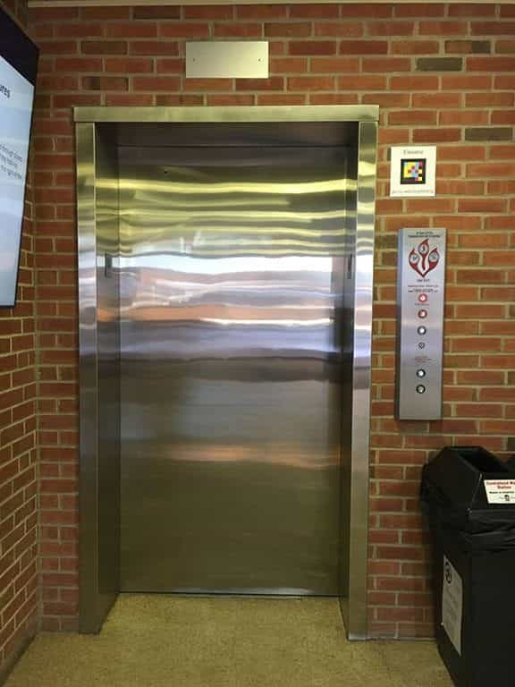 Aufzug in einem NIU-Gebäude mit einem NaviLens-Code mit der Aufschrift „Elevator — go.niu.edu/wayfinding“, der an der Ziegelwand neben der Tastenplatte angebracht ist