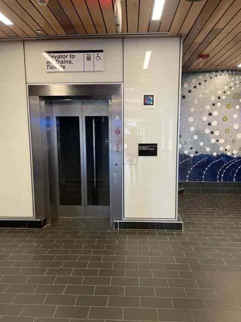Elevator to tracks and ticket office with «Elevator to All Trains, Tickets» sign on the left and a NaviLens code next to the door; on the right, the blue-and-white artistic mosaic typical of White Plains