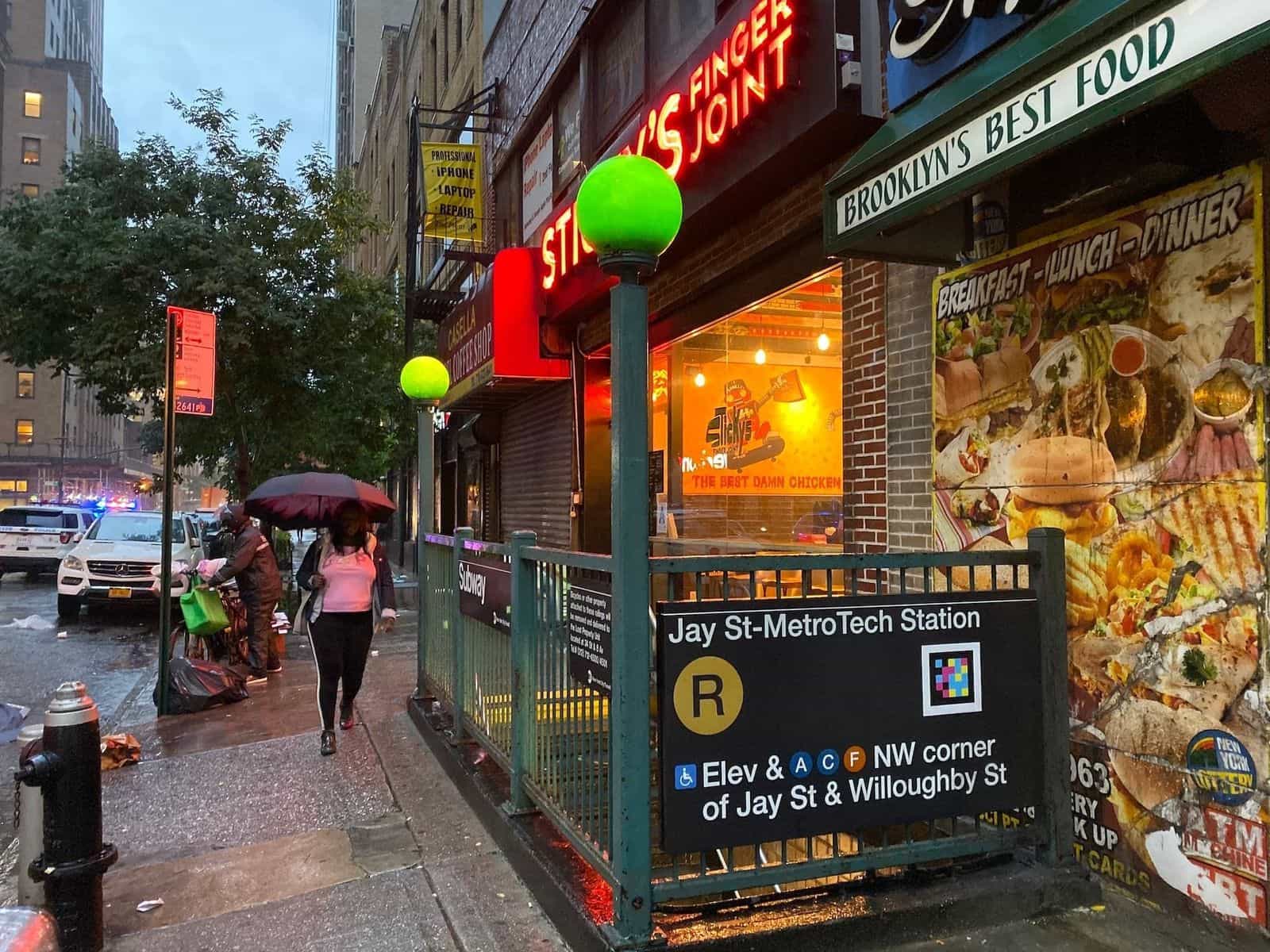 Entrance to Jay St-MetroTech on a rainy day, next to the red neon sign of «Sticky's Finger Joint». On the green railing, the subway sign with the R bullet and a colorful square NaviLens code