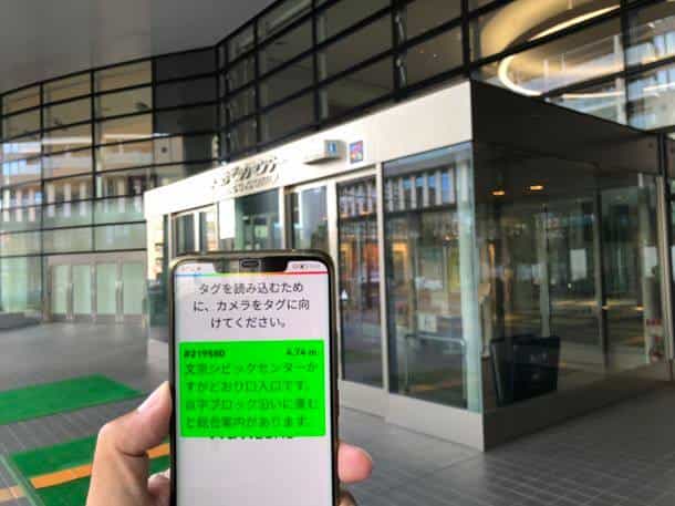 Person scanning the NaviLens code #219580 at the Bunkyo Civic Center entrance; the app announces 4.74 m to the tactile paving guide of the main reception
