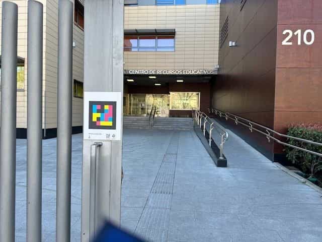 Eingang des ONCE-Bildungsressourcenzentrums in Madrid: Gebäude mit dem Schriftzug „CENTRO DE RECURSOS EDUCATIVOS“ und einer großen 210 an der Fassade; im Vordergrund ein weißer Pfosten mit einem großformatigen farbigen NaviLens-Code und einer barrierefreien Rampe zum Gebäude