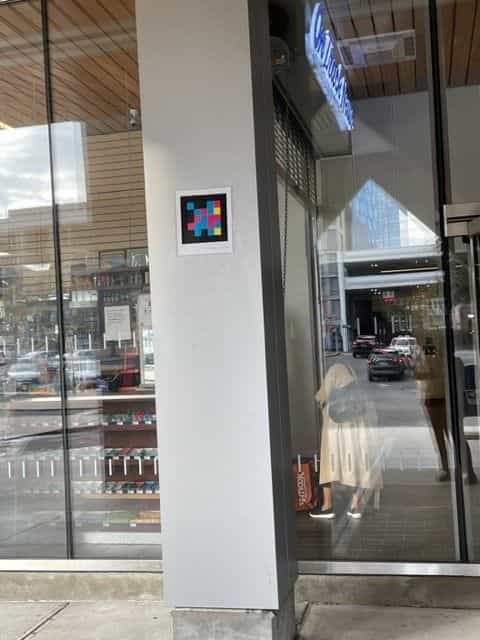 Side entrance to White Plains station: white pillar with a framed NaviLens code, glass storefront and, on the right, an «On Track» illuminated sign; in the background, a traveller in a light dress carrying a Daiso bag