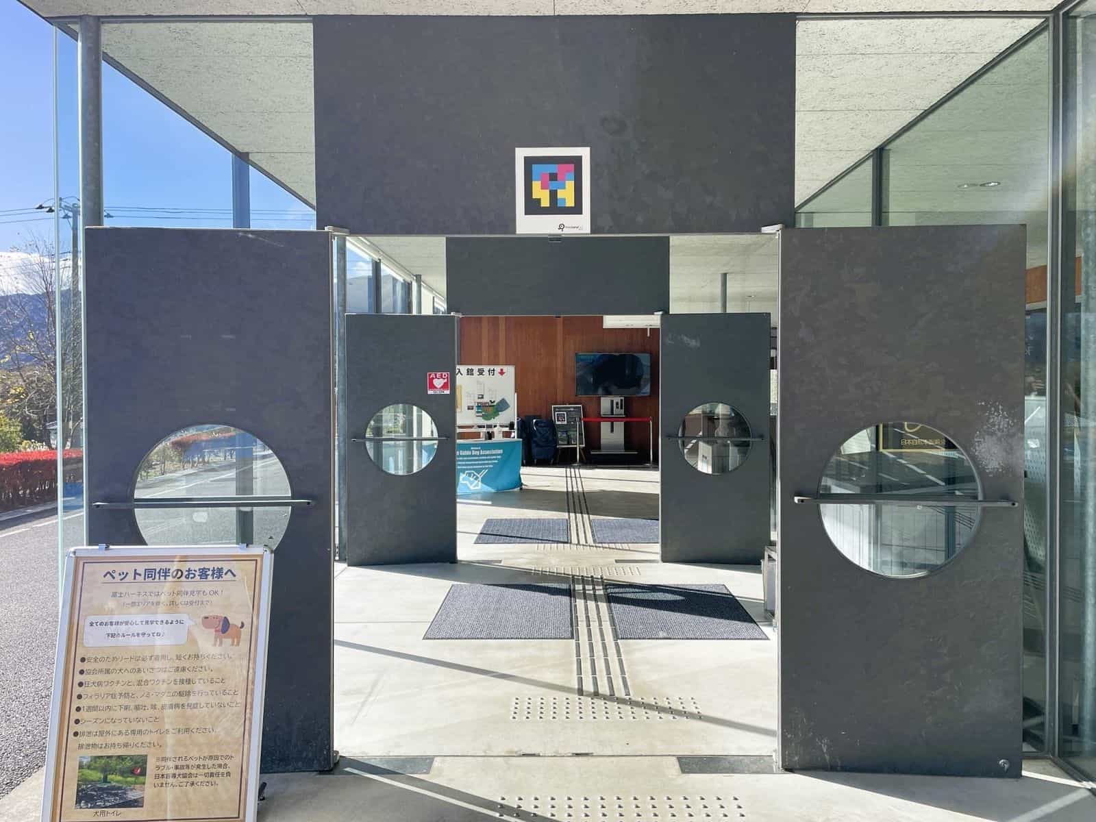 Main entrance of the Japan Guide Dog Center “Fuji Harness”: gray doors with porthole windows, tactile paving leading to the reception desk and, above the lintel, a large NaviLens GO code welcoming visitors