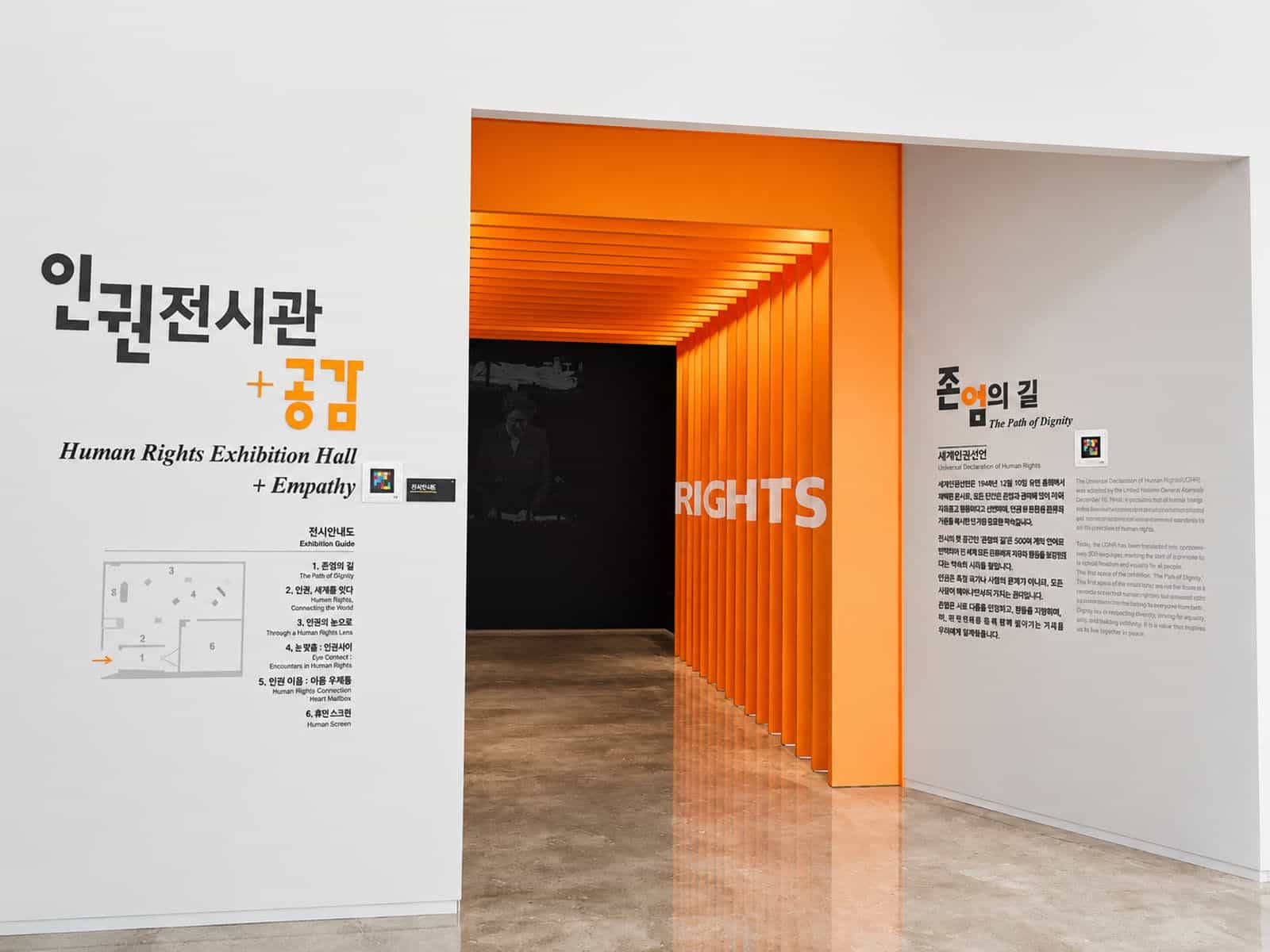 国家人権教育院の「人権展示館＋共感」の入口。韓国語と英語の展示案内図が書かれた白い壁、案内表示の横のNaviLensコード、奥には最初のゾーンのオレンジ色のトンネル