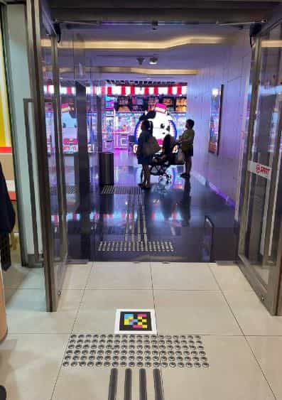 Vista a través de las puertas correderas de cristal del Bukit Panjang Integrated Transport Hub: en primer plano, el suelo con franjas podotácticas grises de botones y, sobre una baldosa cuadrada blanca, un código NaviLens grande en cuadros amarillos, rosas, azules y negros; al fondo, una persona en silla de ruedas eléctrica saliendo del centro comercial Hillion Mall, con escaparates iluminados y una mascota rosa al fondo