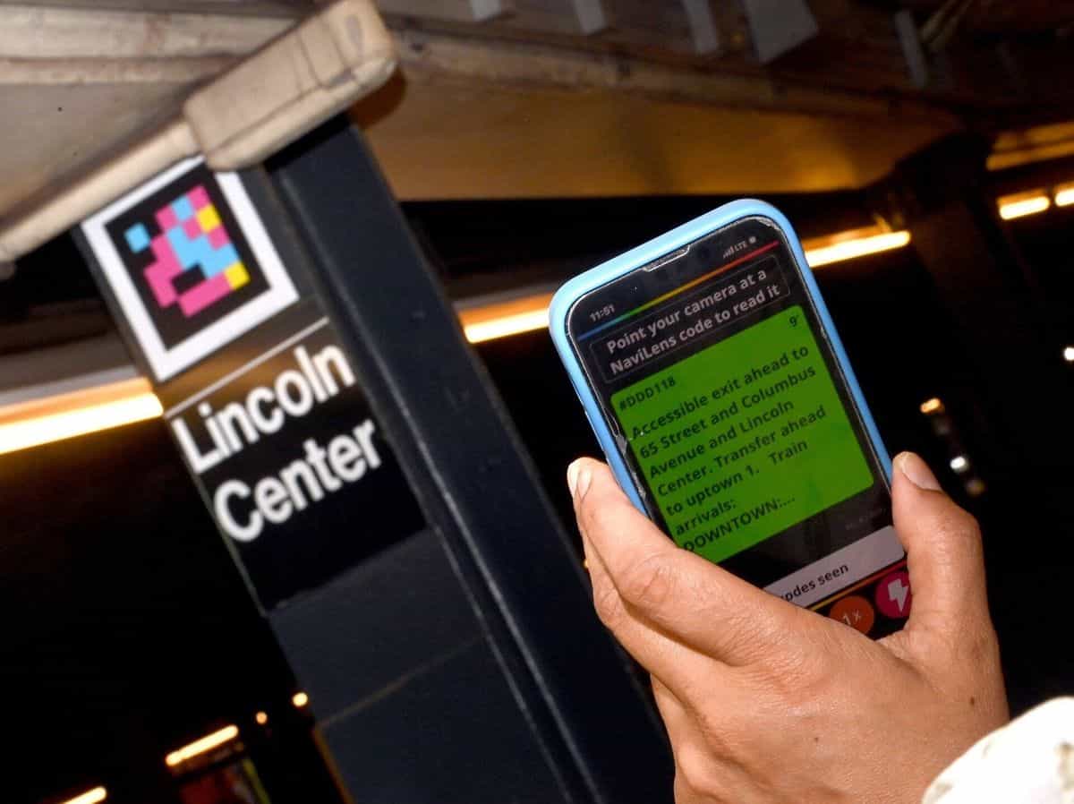 Entrada de la estación 66 St-Lincoln Center (línea 1): columna negra con código NaviLens grande y rótulo «Lincoln Center»; en primer plano, una mano sostiene un móvil con la app NaviLens mostrando en pantalla verde el texto «#DDD118 — Accessible exit ahead to 65 Street and Columbus Avenue and Lincoln Center. Transfer ahead to uptown 1. Train arrivals: DOWNTOWN…»