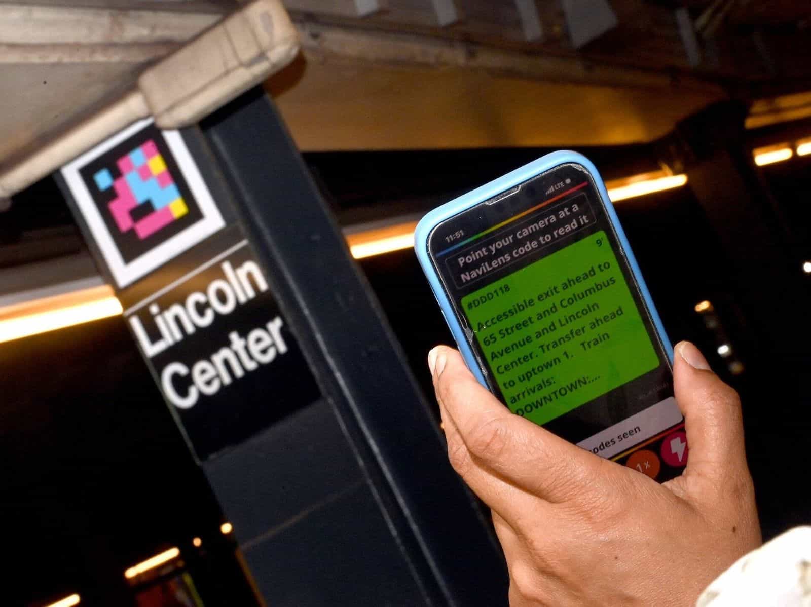 Eingang 66 St-Lincoln Center (Linie 1): schwarze Säule mit großem NaviLens-Code und «Lincoln Center»-Schild; im Vordergrund eine Hand mit Smartphone und der NaviLens-App, auf grünem Bildschirm «#DDD118 — Accessible exit ahead to 65 Street and Columbus Avenue and Lincoln Center. Transfer ahead to uptown 1. Train arrivals: DOWNTOWN…»