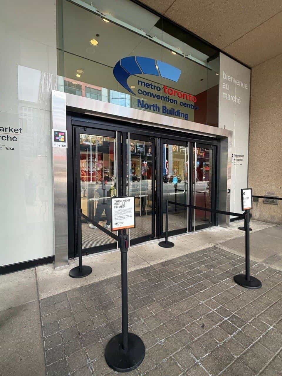 Entrance to the North Building of the Metro Toronto Convention Centre, with the venue sign above the automatic doors and “bienvenue au marché” on the stone wall. On the metal jamb to the left, a NaviLens code with the TIFF logo.