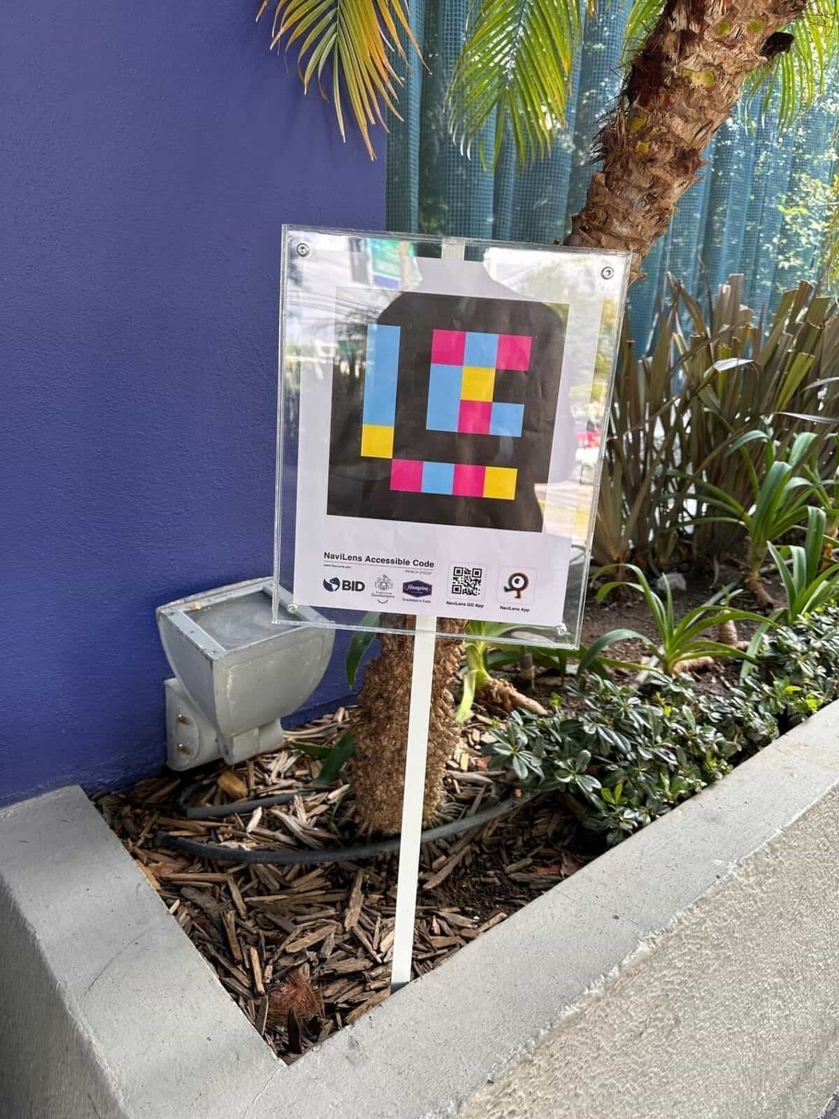 Panneau NaviLens sur un poteau blanc à l'entrée principale de l'Hampton, à côté du jardin et de la façade bleue