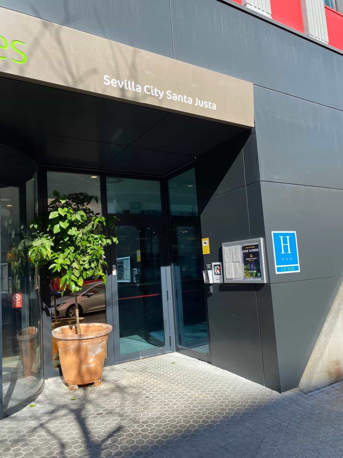 Haupteingang des ibis Styles Sevilla City Santa Justa: Glasdrehtür mit Schild „Sevilla City Santa Justa“, Orangenbaum im Tontopf, blaues „H Ciudad“-Piktogramm und NaviLens-Code an der Tür
