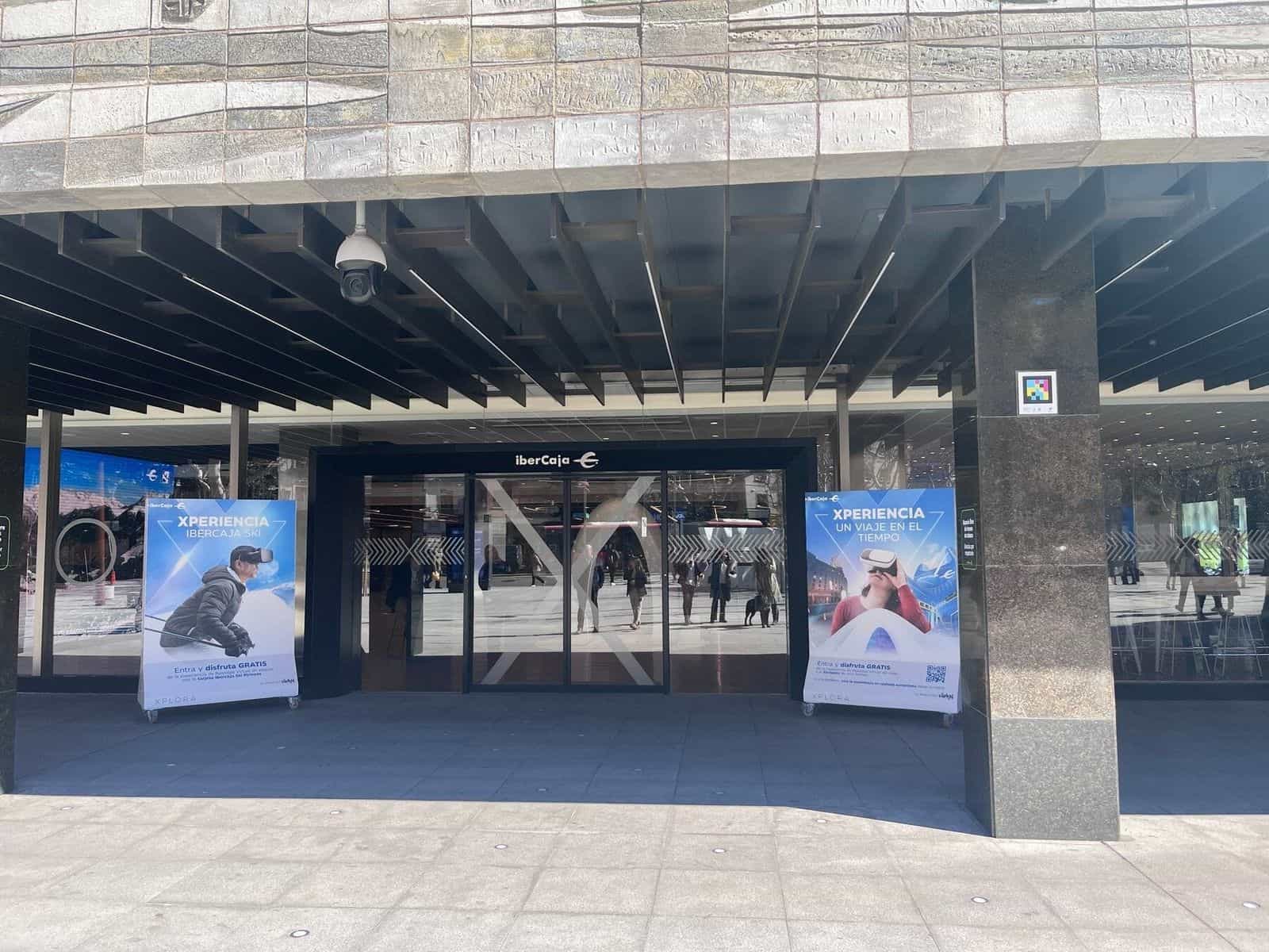 Acceso principal de Ibercaja en Zaragoza con dos carteles publicitarios «Xperiencia Ibercaja Ski» y «Un viaje en el tiempo» y un código NaviLens pegado en la columna de granito negro