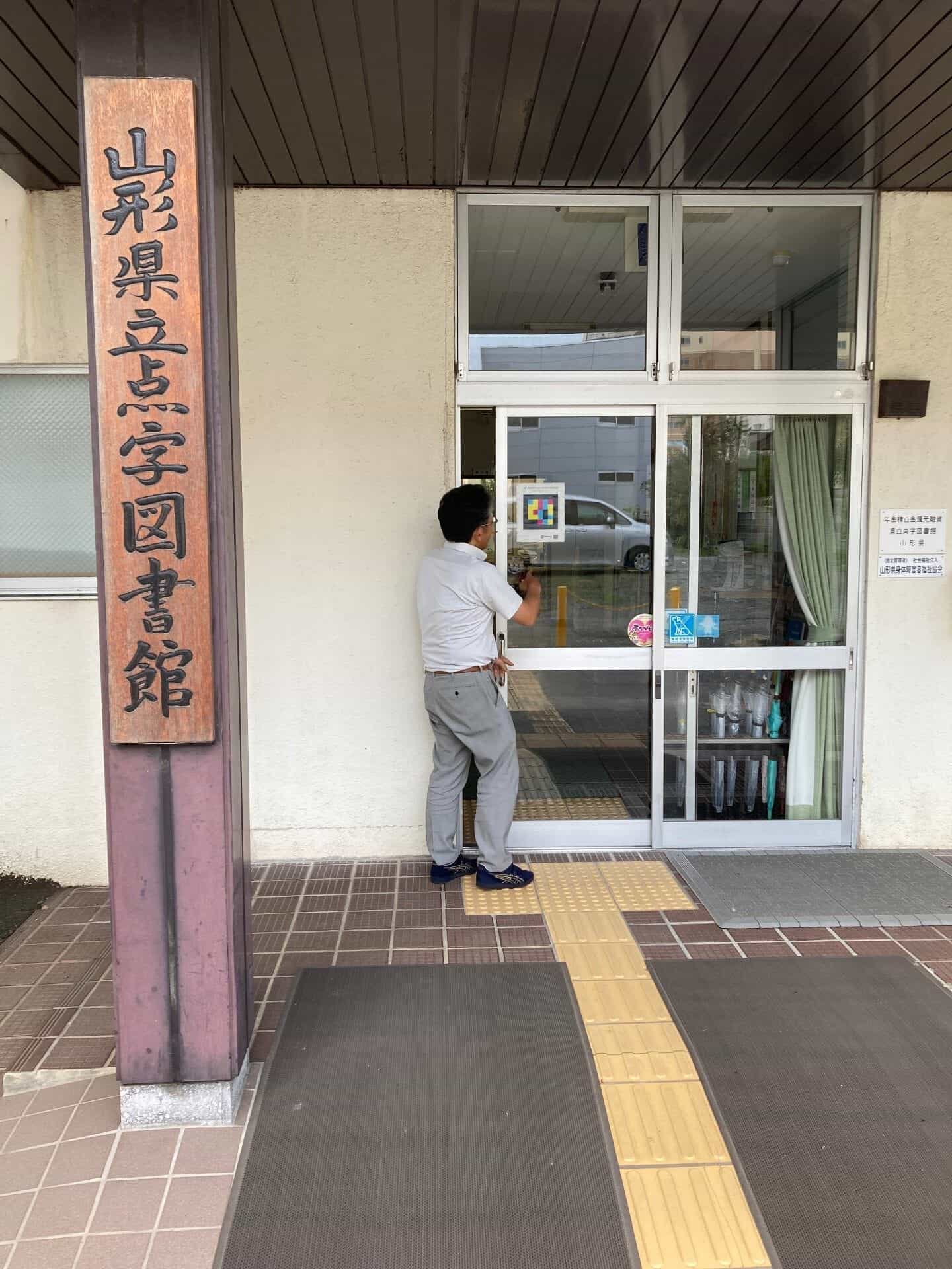 山形市にある山形県立点字図書館のメインエントランス。大きな木製の縦型看板には「山形県立点字図書館」の漢字が彫られ、床には黄色の点字ブロックが敷かれている。白いポロシャツを着た職員が自動ガラスドアを開けており、そのドアには「Empowering visually impaired」というスローガンが書かれたカラフルな四角いNaviLensコードが貼られている