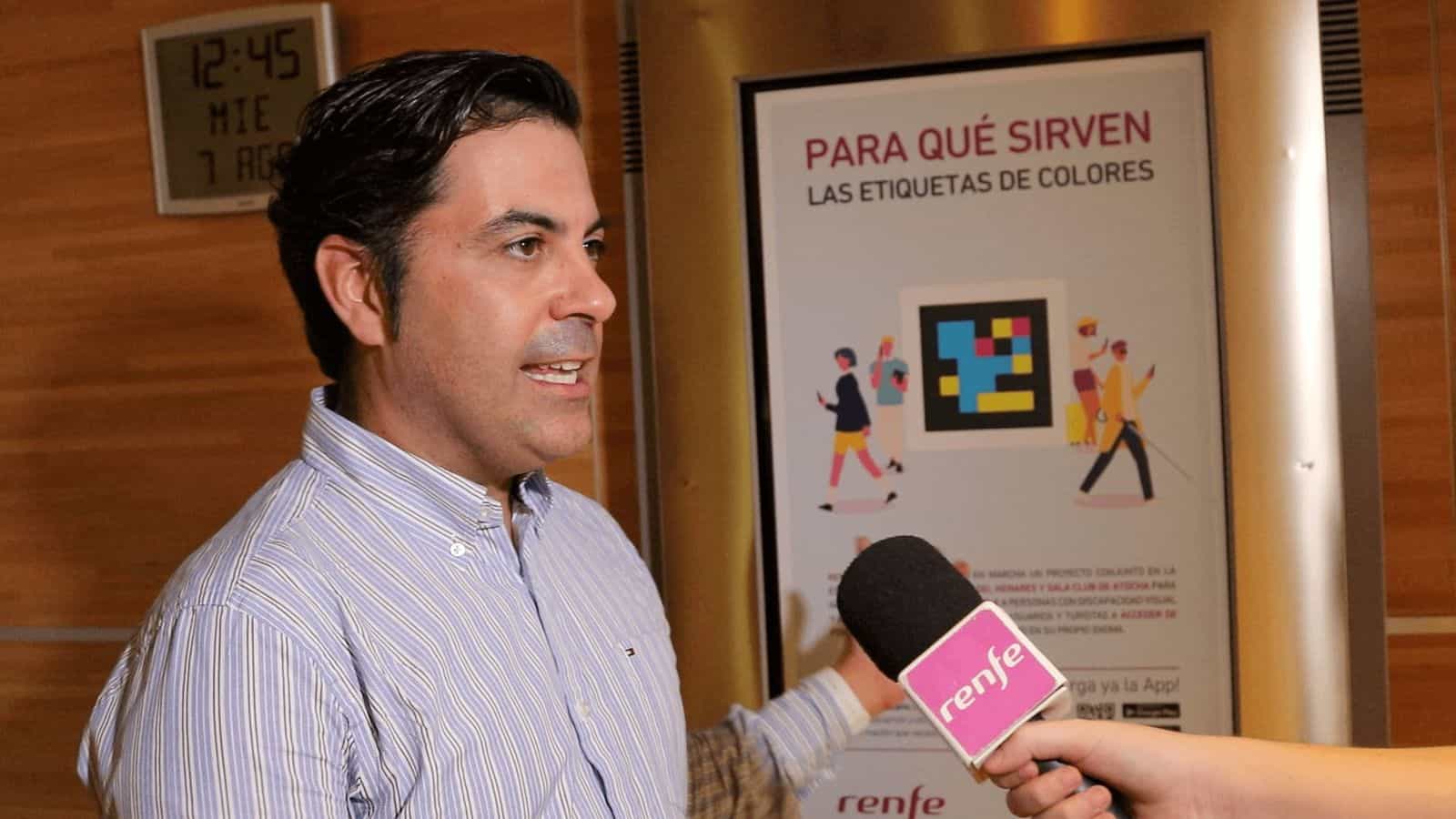 A Renfe spokesperson addresses the press in front of a poster reading ‘What the color tags are for' with a large NaviLens code, during the Atocha pilot launch; in the foreground, a microphone with the Renfe logo.