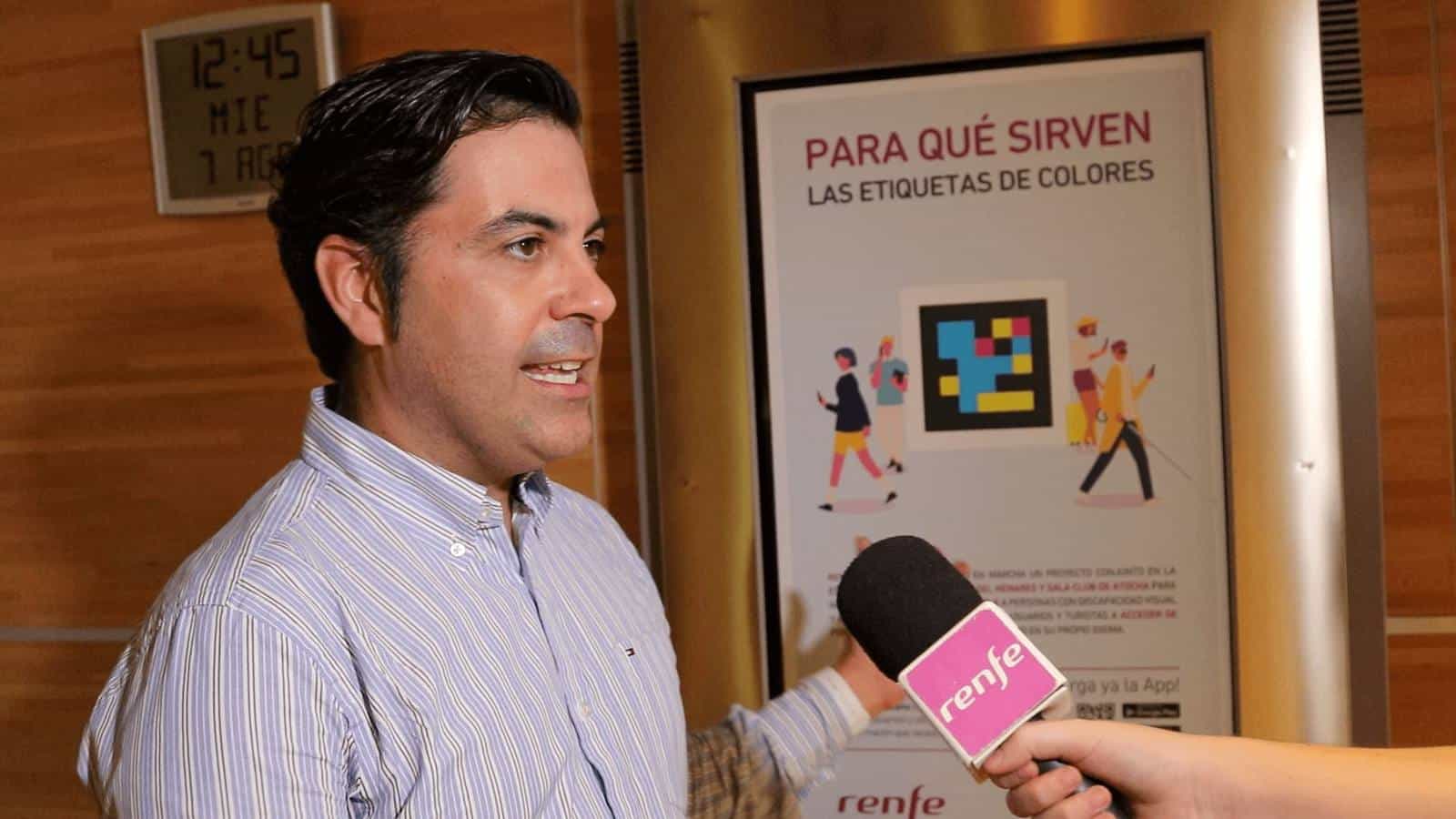 A Renfe spokesperson addresses the press in front of a poster reading ‘What the colour tags are for' with a large NaviLens code, during the Atocha pilot launch; in the foreground, a microphone with the Renfe logo.