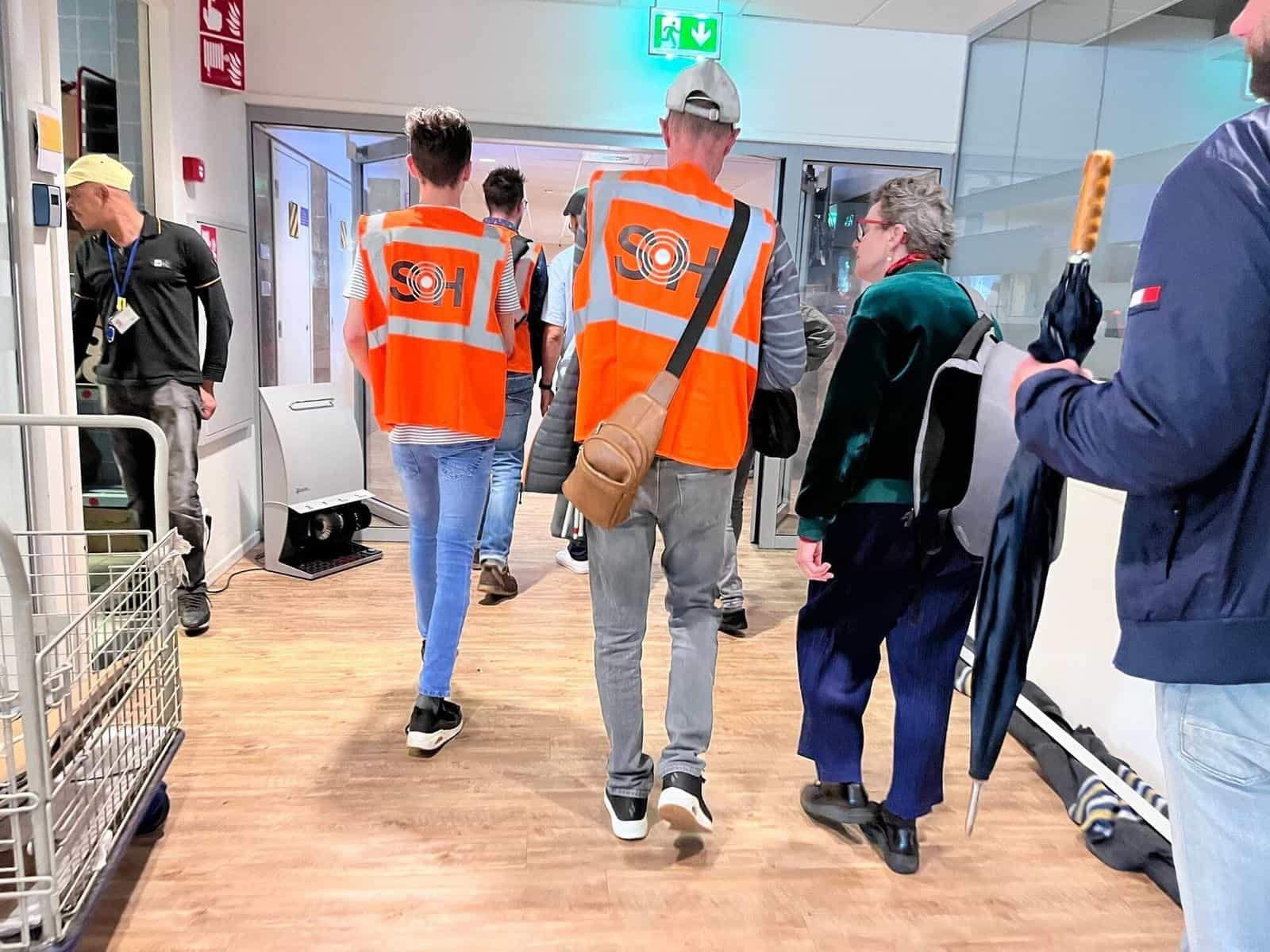 Squadra del test di NaviLens a Den Haag Centraal —gilet arancioni con il logo SH (Stay Healthy)— che accompagna una persona utente in un corridoio interno della stazione durante la giornata di valutazione