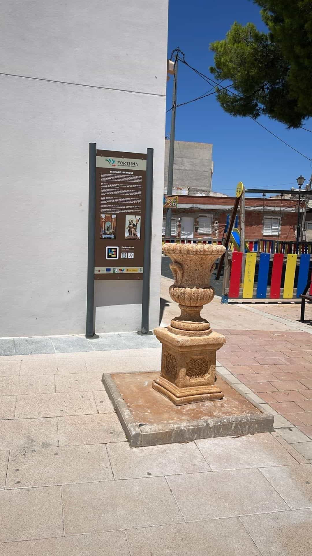 Ermita de San Roque (Fortuna): tótem patrimonial junto a una jarra de piedra y a una zona de juegos infantil, con código NaviLens escaneable y cartela del templo del siglo XVIII