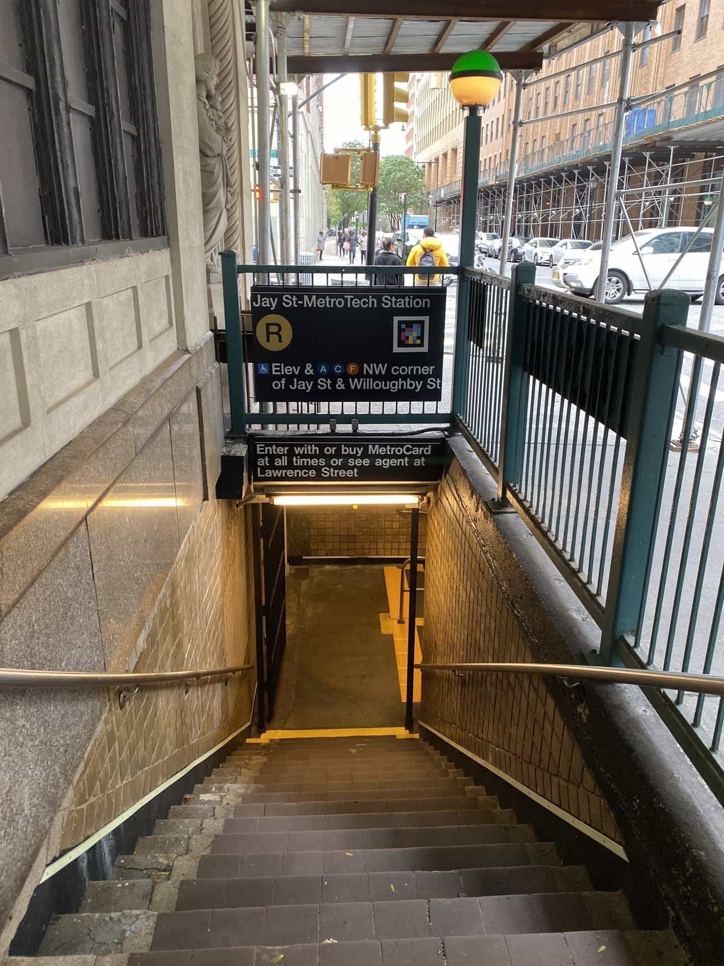 Another street entrance to Jay St-MetroTech under construction scaffolding: green railing, the classic NYC subway green globe and, on the black «Jay St-MetroTech Station» sign, the R bullet and a NaviLens code next to the message «Elev & A C F NW corner of Jay St & Willoughby St»
