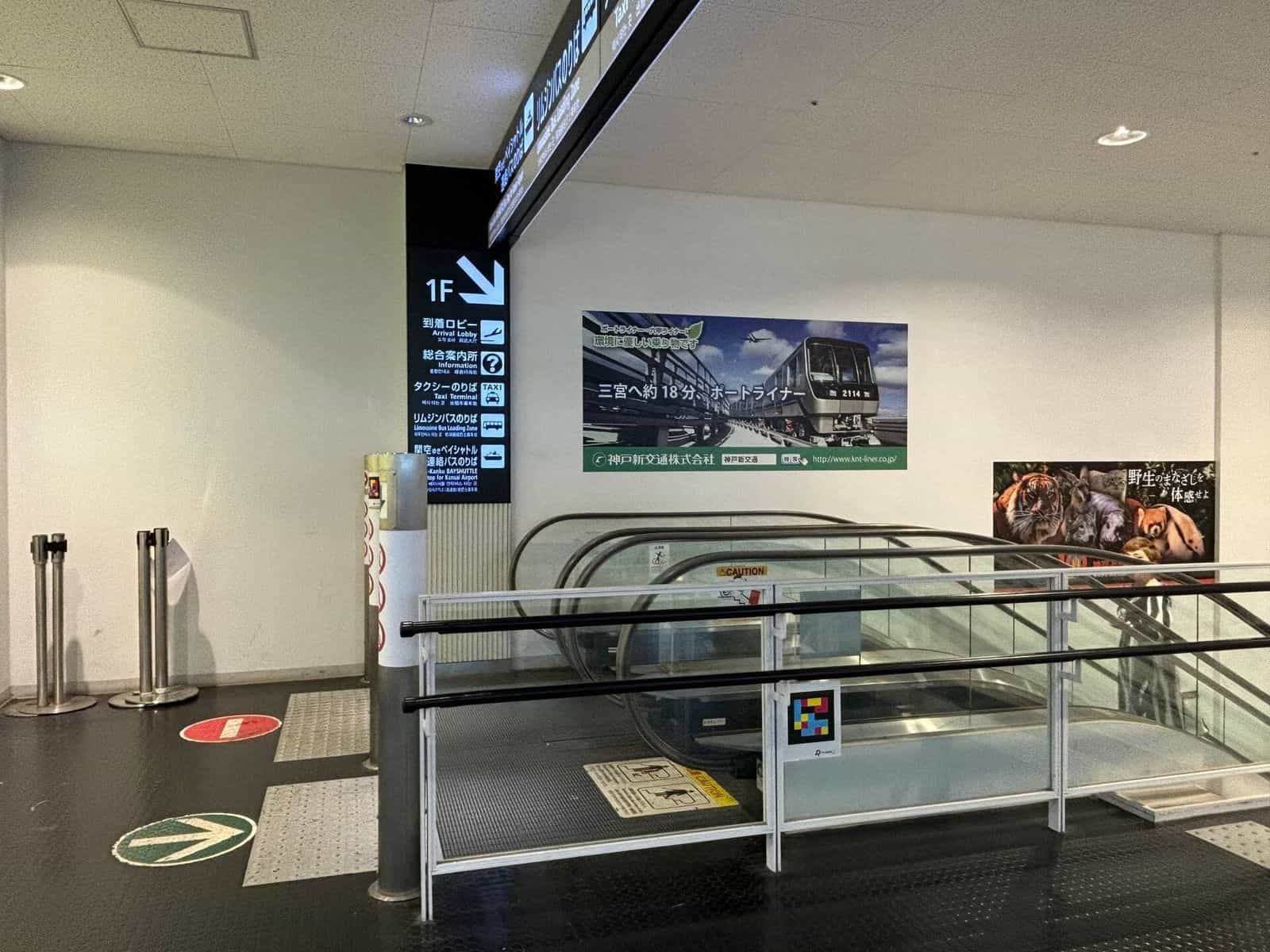Rolltreppe abwärts zum 1F im Kobe Airport mit schwarzem Schild «1F · Arrival Lobby» und NaviLens-Tag am Handlauf-Glas