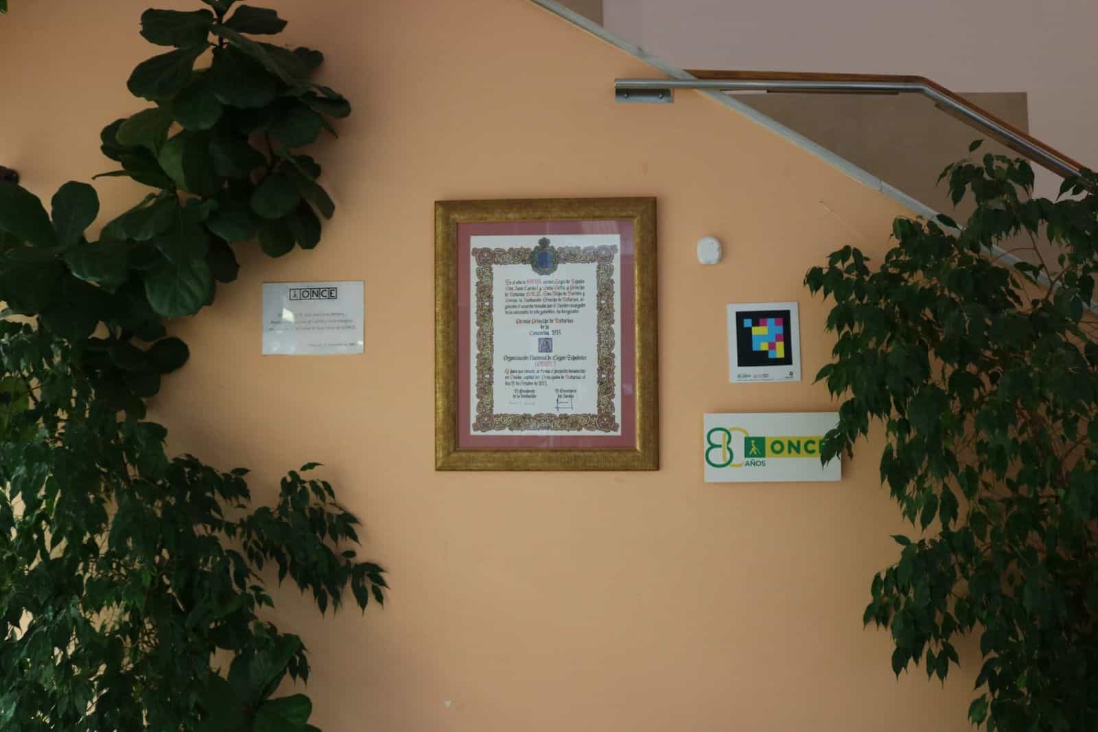 Vano scale dell’ONCE Valladolid: parete color salmone con una targa incorniciata del Premio Príncipe de Asturias de la Concordia 2013 assegnato all’ONCE; a destra, un codice NaviLens quadrato a colori e sotto il logo commemorativo «80 años — ONCE», inquadrato da foglie di ficus
