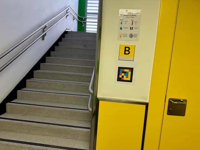 Staircase at CRE ONCE Madrid, zone B: metal handrail, contrast-marked steps and, on the yellow landing wall, a “B” sign and a square colour NaviLens code below it