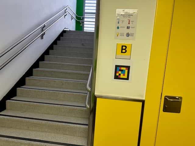 Treppenhaus im CRE ONCE Madrid, Zone B: Metallgeländer, kontrastmarkierte Stufen und an der gelben Treppenabsatzwand ein „B“-Schild und darunter ein quadratischer NaviLens-Farbcode