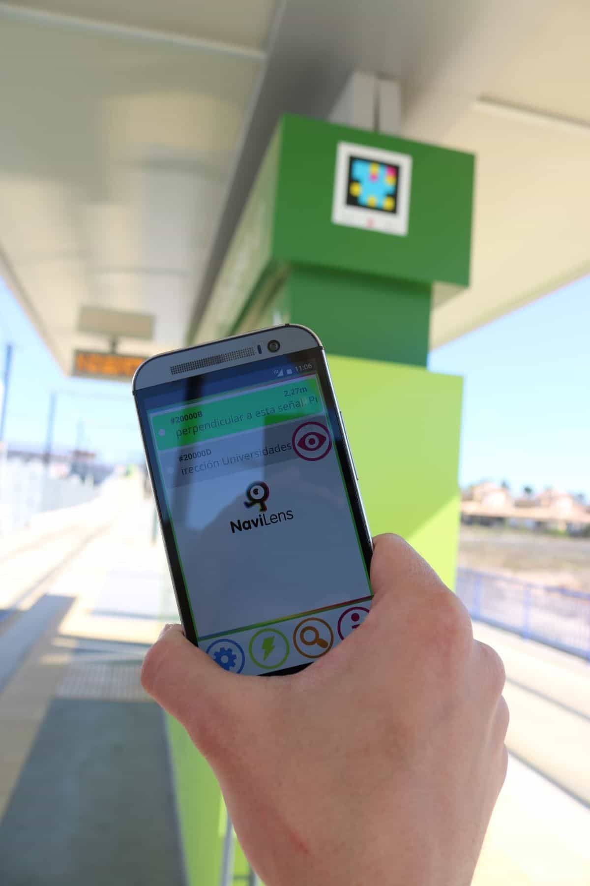 Persona escaneando un código NaviLens situado en lo alto de una marquesina del Tranvía de Murcia, con la pantalla LED de próximas llegadas al fondo