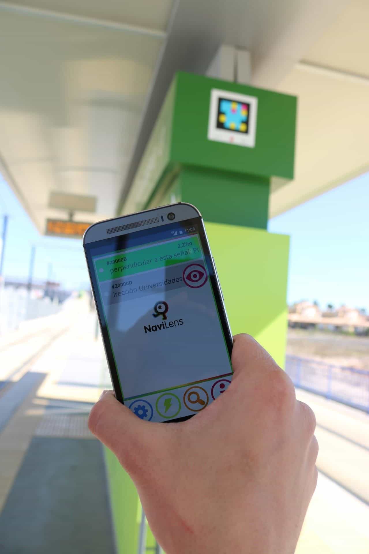 Persona che inquadra un codice NaviLens collocato in cima a una pensilina del Tranvía de Murcia, con lo schermo LED dei prossimi arrivi in fondo