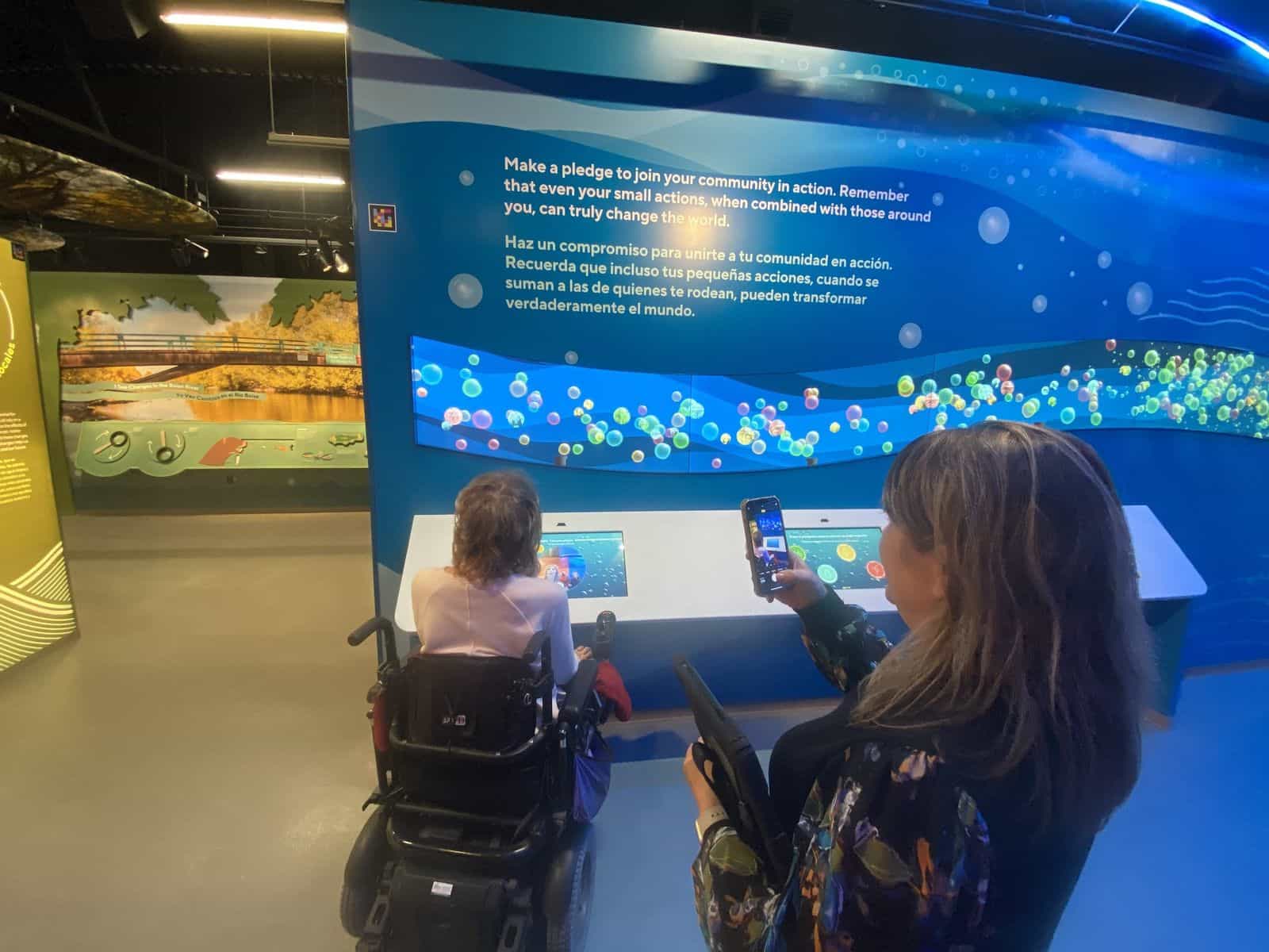 Zwei Besucherinnen an der Pledge Wall von The WaterShed, unter dem zweisprachigen Text „Make a pledge to join your community in action / Haz un compromiso para unirte a tu comunidad en acción“: eine Rollstuhlnutzerin bedient den Touchscreen, während eine zweite Besucherin ihr Handy hochhält, auf dessen Display die NaviLens-App den Code an der Säulenkante liest