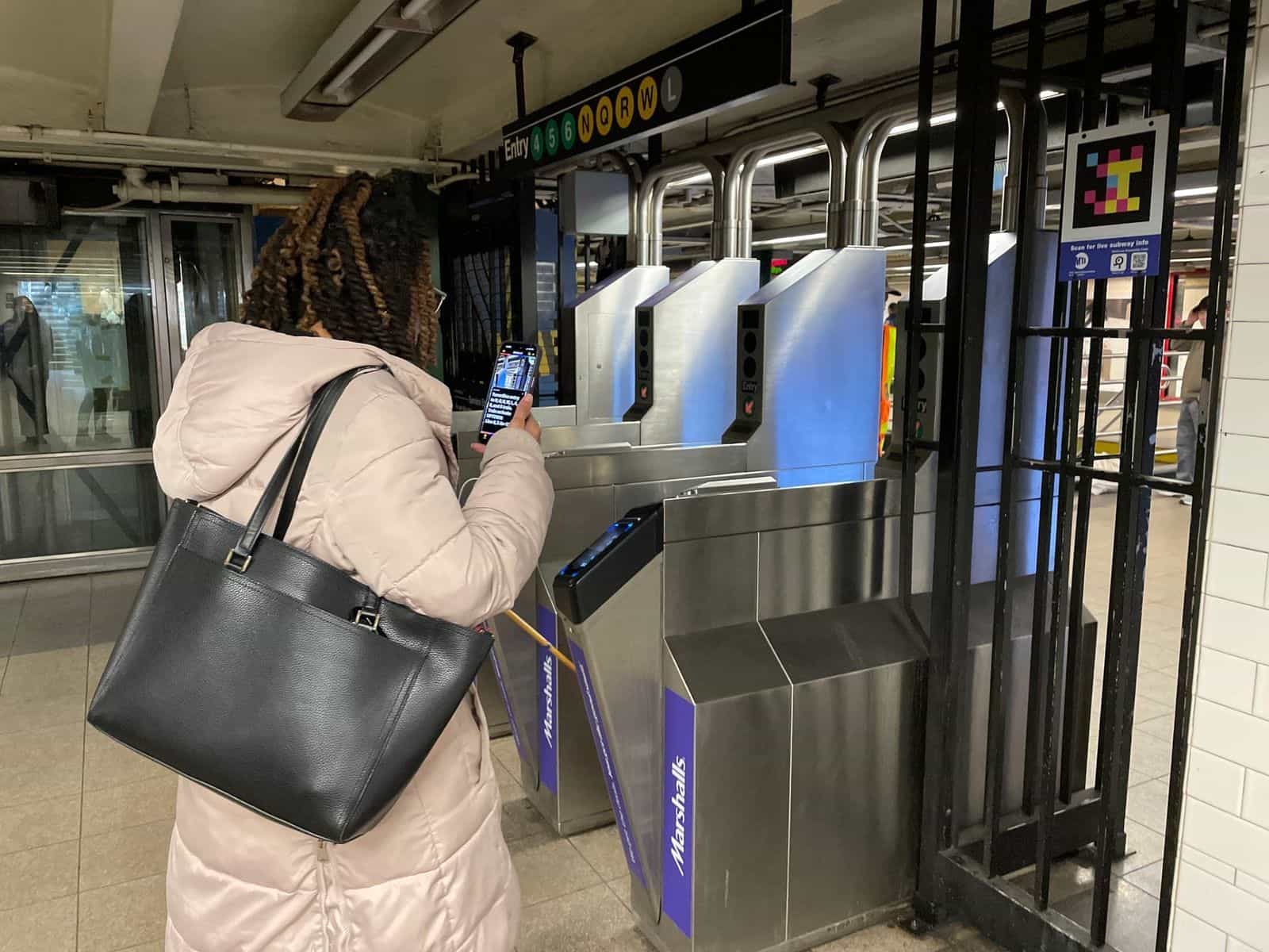 User with white cane scans a NaviLens code high on the turnstile partition with their phone; on screen, a long wayfinding text generated by the app
