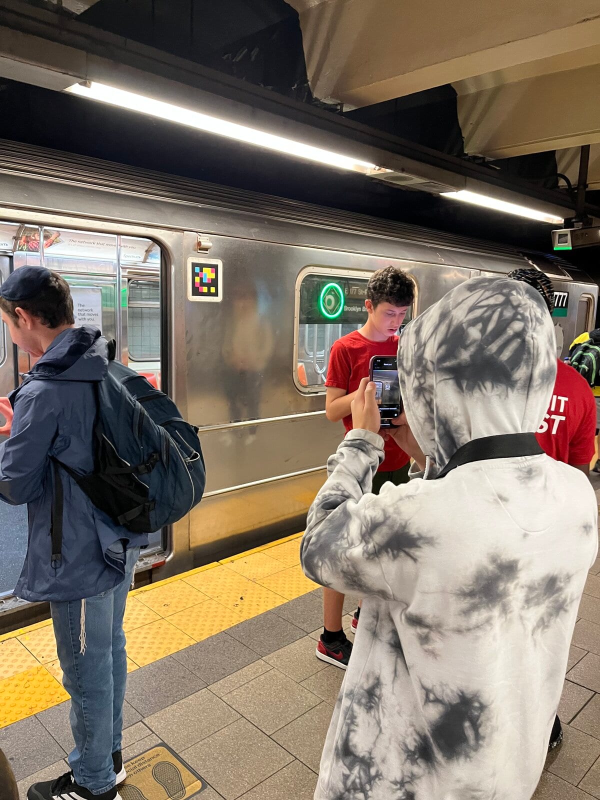 Membre de l'équipe NaviLens sur un quai du métro de NY scannant avec son téléphone un code NaviLens collé sur le côté de la voiture ; à côté de la fenêtre éclairée on lit «E · 179 St-Brooklyn»