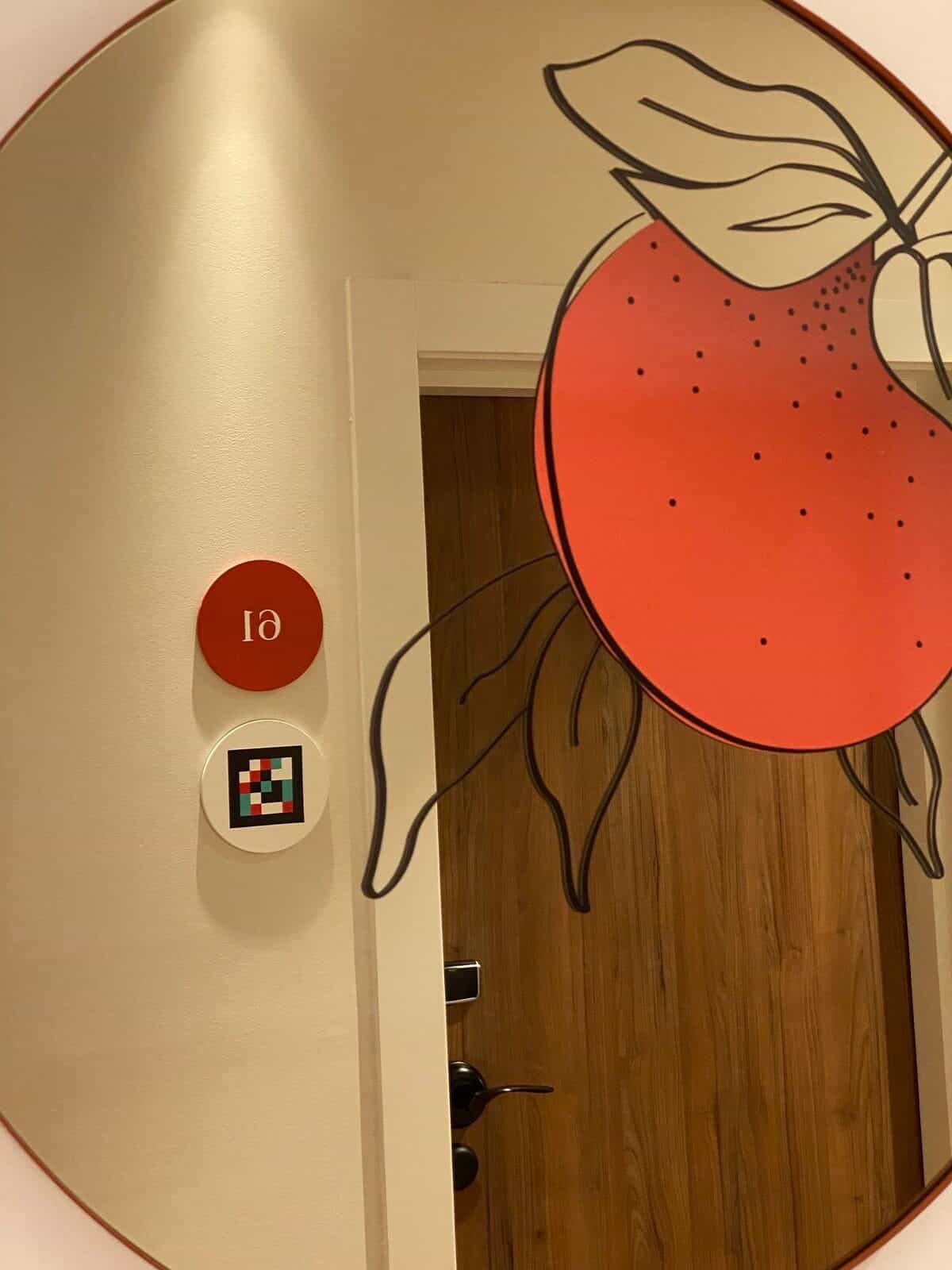 Flur am ibis Styles Sevilla im großen runden Spiegel mit rotem Rahmen reflektiert: oranger „61“-Disc spiegelverkehrt und NaviLens-Code darunter
