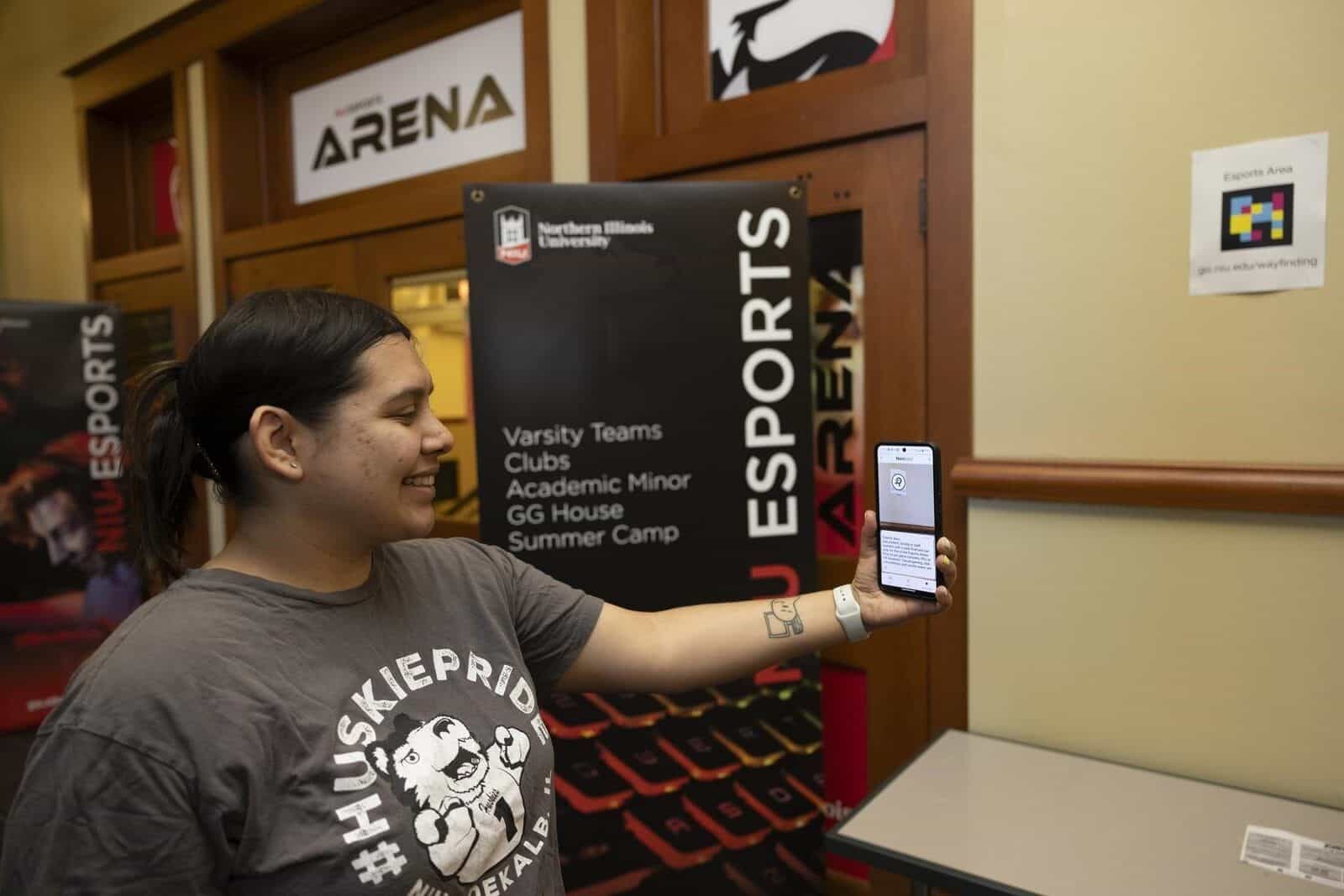 Student consulting a NaviLens code with their mobile phone next to the NIU Esports Arena sign (Varsity Teams, Clubs, GG House, Summer Camp)