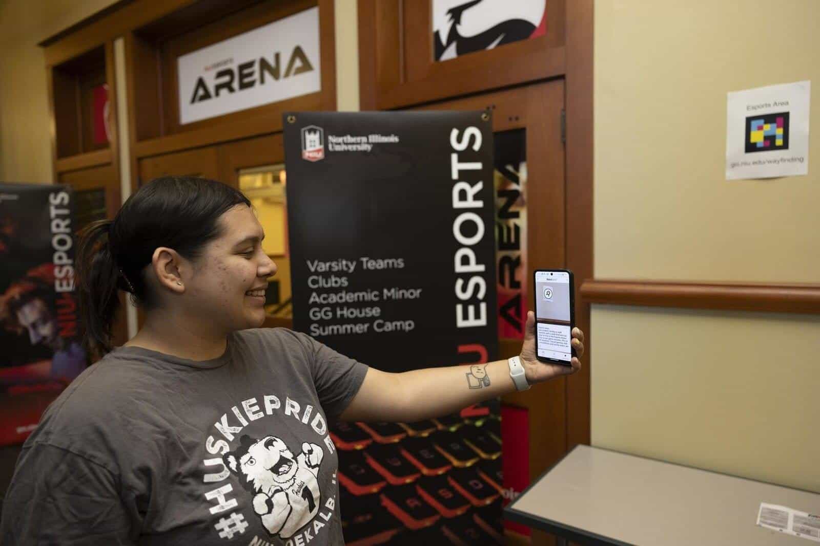 Student consulting a NaviLens code with their mobile phone next to the NIU Esports Arena sign (Varsity Teams, Clubs, GG House, Summer Camp)