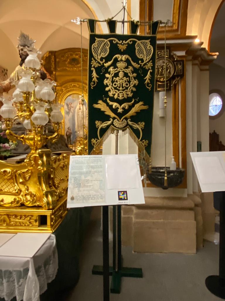 Green banner embroidered in gold of the Brotherhood of the Christ of Hope next to a lectern with the accessible caption and the NaviLens code