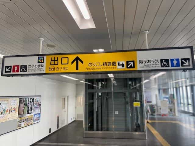 神戸空港駅の黒地に黄色の吊り下げサイン。3つの絵文字:NaviLensタグ付きの「女子お手洗」、「出口 ↑」と「のりこし精算機」、NaviLensタグ付きの「男子お手洗」