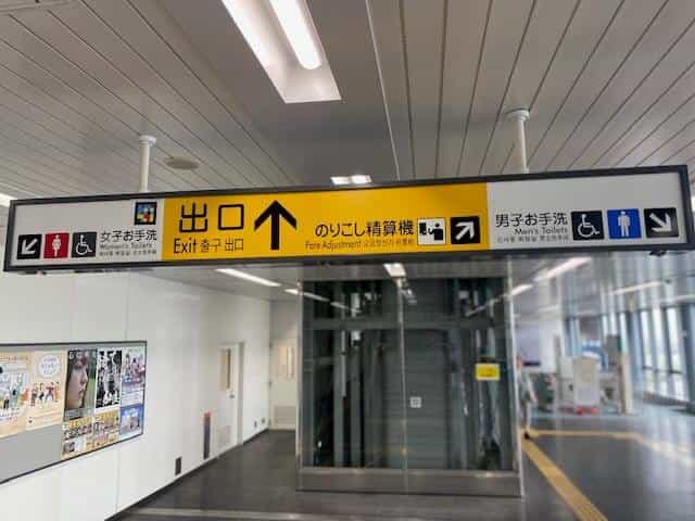 Black-and-yellow hanging sign at Kobe Airport Station with three pictograms: “Women's Toilets” with NaviLens tag, “Exit ↑” and “Fare Adjustment”, and “Men's Toilets” with NaviLens tag