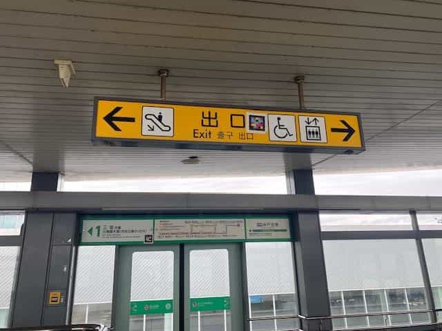 神戸空港駅の黄色い吊り下げサイン「出口 / Exit / 출구」とエスカレーター、エレベーター、車いす絵文字、左右の矢印、絵文字の中央に配置されたNaviLensタグ