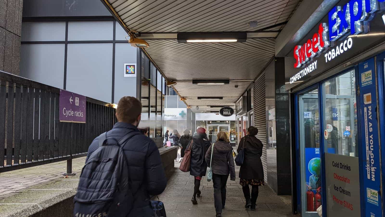 Covered pedestrian access to Euston Station with shops (Sweet Express, Pharmacy, William Hill, WHSmith) and NaviLens code visible on the metallic wall