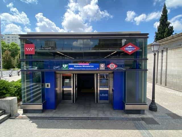 Frontansicht des Zugangs Nuevos Ministerios von der Straße: Glaskubus mit blauer Struktur, oben die rote Metro-Raute, die rote Flagge mit vier weißen Sternen der Comunidad de Madrid, das blaue Schild „Nuevos Ministerios“ mit den Symbolen der Linien 6, 8 und 10 und ein NaviLens-Code am Identifikationsstreifen neben dem Haupteingang