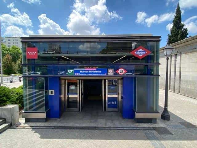 Front view of the Nuevos Ministerios access from the street: glass cube with blue structure, the Metro red diamond on top, the red flag with four white stars of the Community of Madrid, the blue “Nuevos Ministerios” sign with the icons of lines 6, 8 and 10, and a NaviLens code on the identification strip next to the main entrance