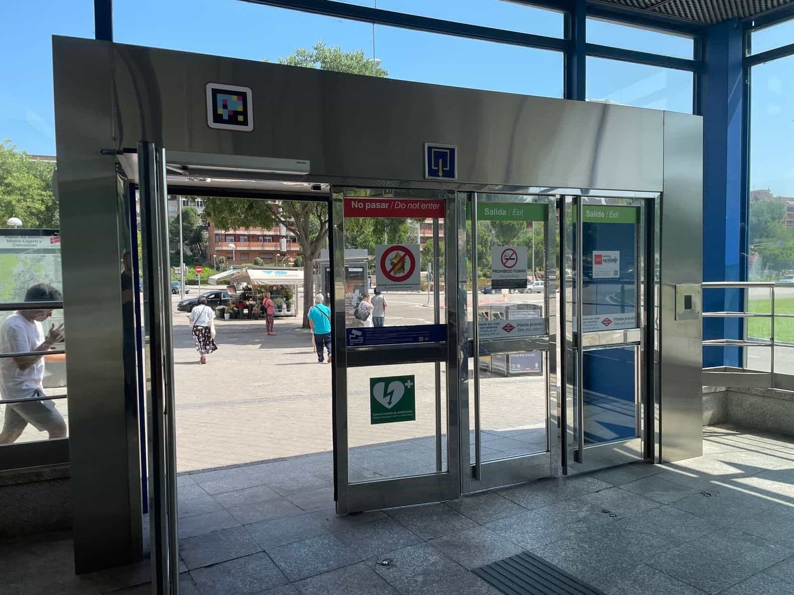 Außenzugang Mar de Cristal: der Glas- und Edelstahlkubus der Station mit einem weiß gerahmten NaviLens-Code am Metallsturz; darunter Schiebetüren mit grünen „Ausgang/Exit“-Streifen, ein grünes Defibrillator-Schild und der Ausgang zur baumbestandenen Allee mit einer Blumenhandlung im Hintergrund