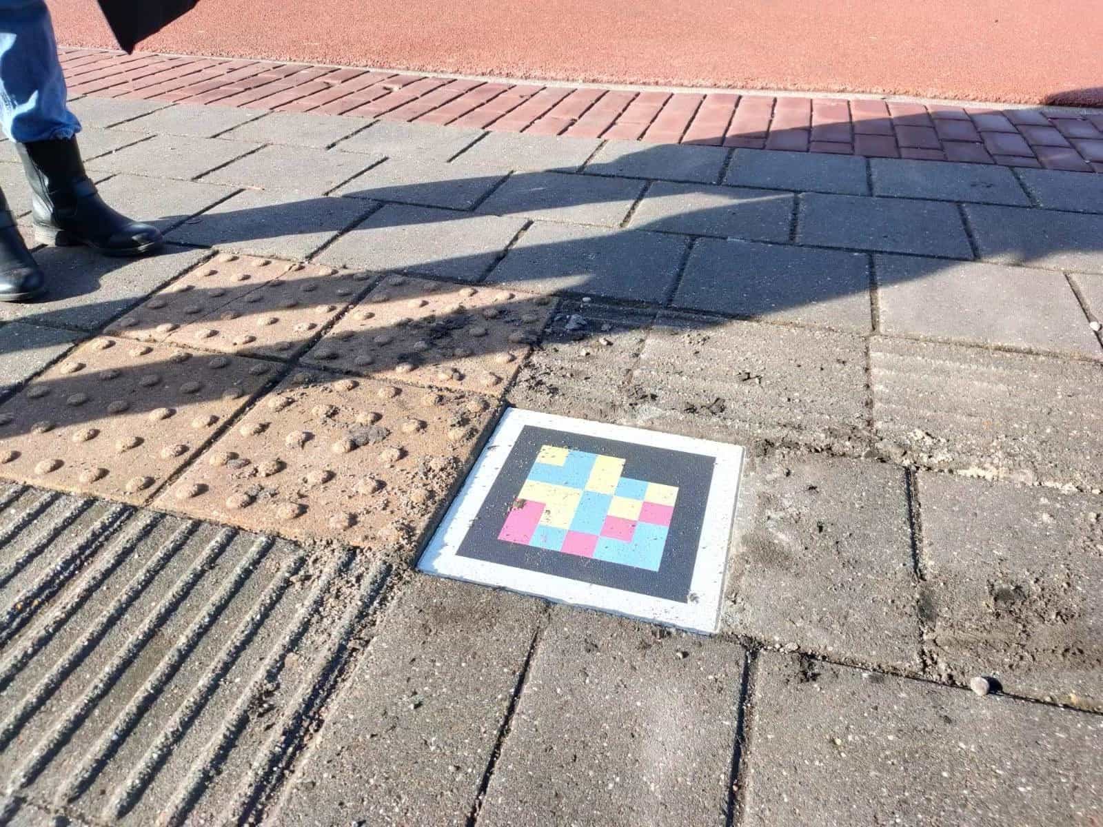 Zugangsweg zum Radboudumc mit taktilen Bodenplatten aus Noppen (Aufmerksamkeitssignal) und Rillen (Richtungsführung). Neben der Noppenplatte ein quadratischer NaviLens-Code bündig im Pflaster, der die Querung und den Klinikeingang ankündigt