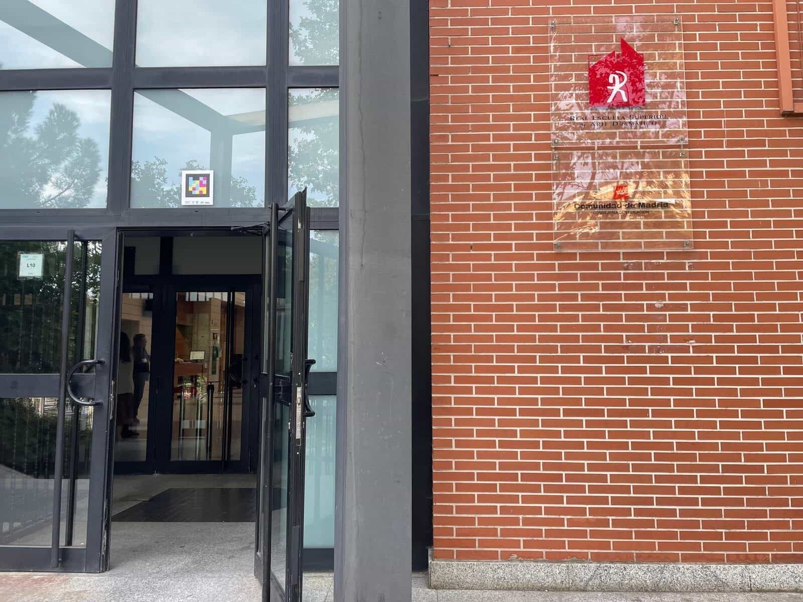 Haupteingang der RESAD: geöffnete Glas- und Stahltüren mit einem weiß gerahmten NaviLens-Code auf dem Glas des Eingangswürfels; rechts auf der roten Backsteinmauer die institutionelle Tafel mit dem roten Logo der RESAD und dem Wappen der Comunidad de Madrid · Bildungsministerium