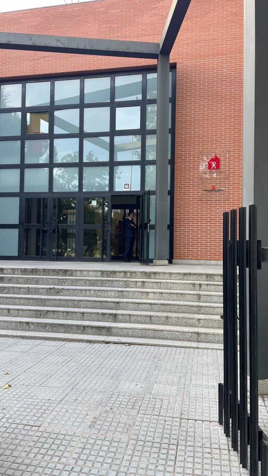 Außenansicht der RESAD von der Straße: große rote Backsteinfassade mit geneigtem Dach und einem verglasten Würfel mit schwarzer Metallkonstruktion; in der Mitte der Glasfront ein weiß gerahmter NaviLens-Code über der geöffneten Tür, an der ein uniformierter Sicherheitsmitarbeiter wartet; im Vordergrund Granittreppen und ein Metallgitter