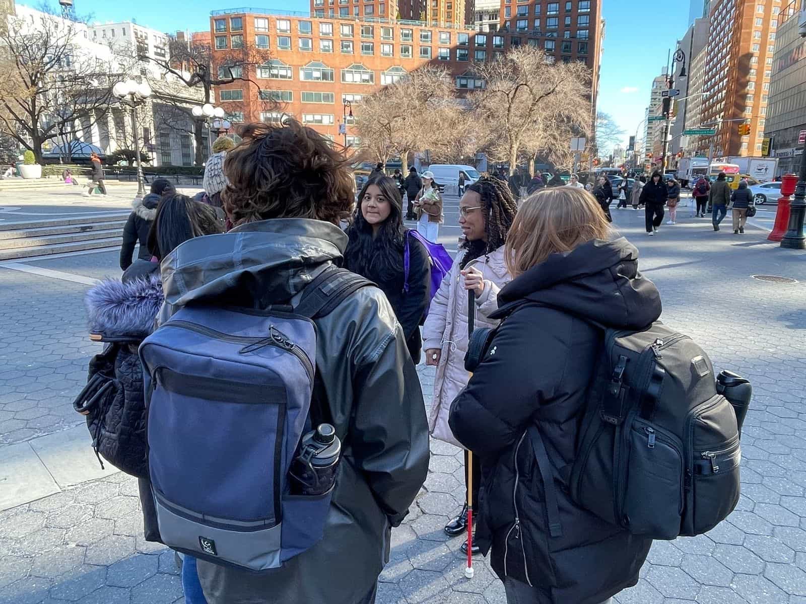 Group of visitors with white canes and backpacks gathered on Union Square plaza; in the background, the stairs down to the subway entrance and the surrounding brick buildings