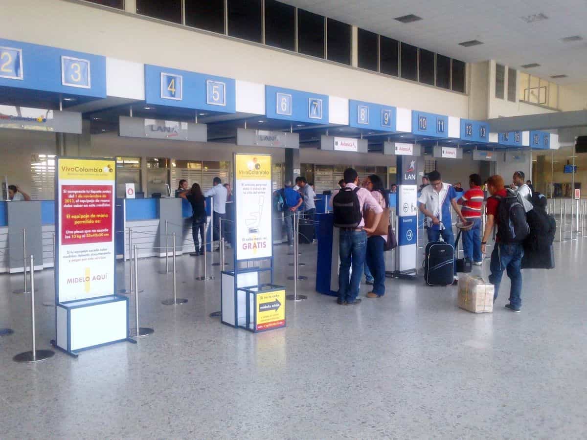 Außenansicht des Flughafens Los Garzones in Montería von der Rollbahn-Anflugzone aus: einstöckiges Terminalgebäude mit weißem Dach, Gärten und Palmen rundum, kolumbianische Flagge am Haupteingang